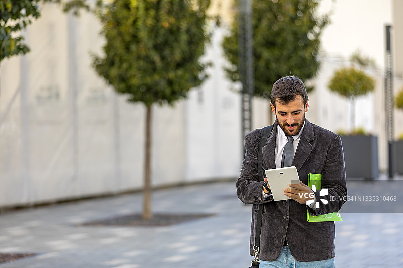 一位年轻的商人正手持电子平板电脑走在城市的街道上。图片素材