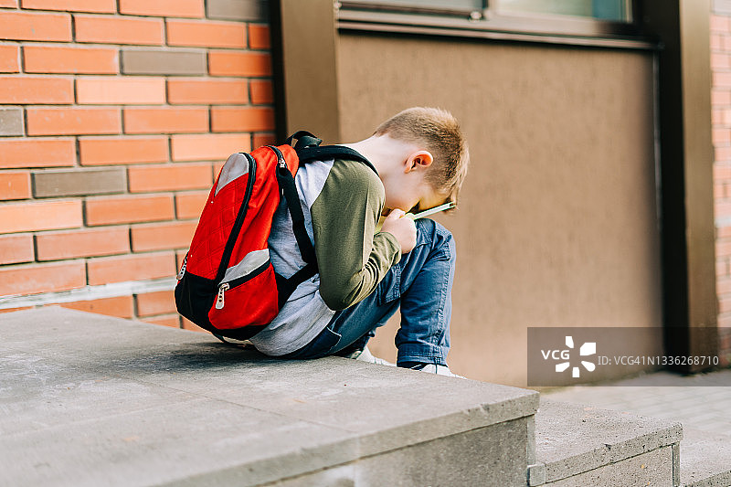 回到学校。可爱的孩子背着书包，拿着记事本和训练书。一名小学生带着书包。小学生去上课。累的孩子坐在楼梯外面的砖墙图片素材