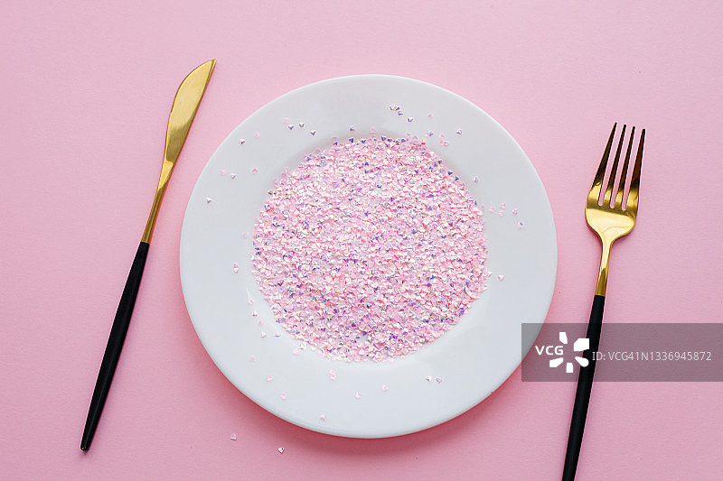 白色的盘子和粉红色的闪光纸屑，金色和黑色的刀叉在粉红色的背景上。营养和节食的概念。图片素材