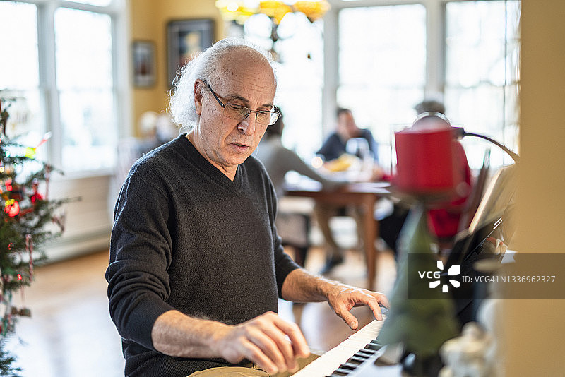 这位72岁的老人，满头银发，是一位音乐家，正在为他的家人弹奏钢琴，他的家人正在明亮宽敞的客厅里用餐，这间客厅是为圣诞节装饰的。图片素材