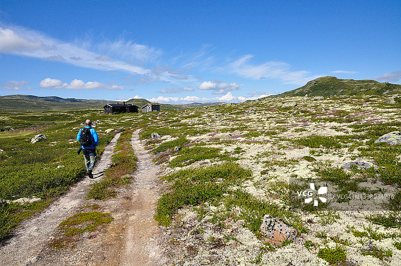 背着背包，穿着蓝色夹克的男人走在通往山间小屋的小径上。在一个美丽的夏末的一天，高山高原的风景。图片素材