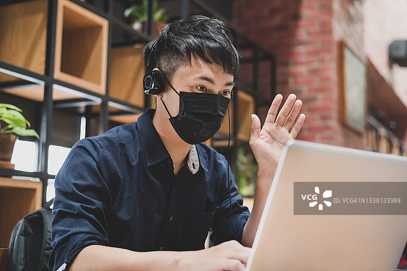 年轻的亚洲华人企业家戴着防护口罩，在办公室里用笔记本电脑与商业伙伴和客户进行视频会议，向他们挥手致意图片素材