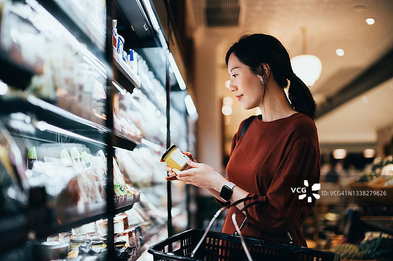 一个年轻的亚洲女人提着一个购物篮，站在牛奶货架上，阅读新鲜有机健康酸奶瓶子上的营养标签。选择更健康的食物图片素材