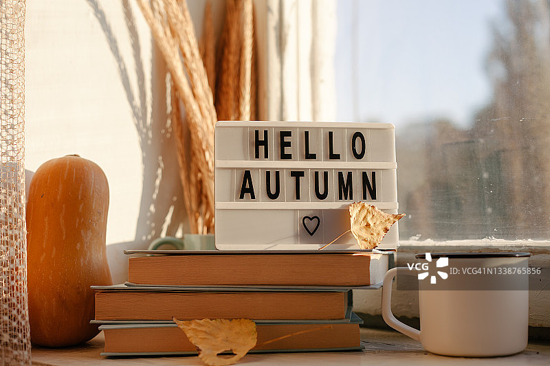 秋天的贺卡。窗台上印有“你好，秋天”字样的灯箱。秋天的水果，苹果和南瓜，干花，书和一杯咖啡。图片素材