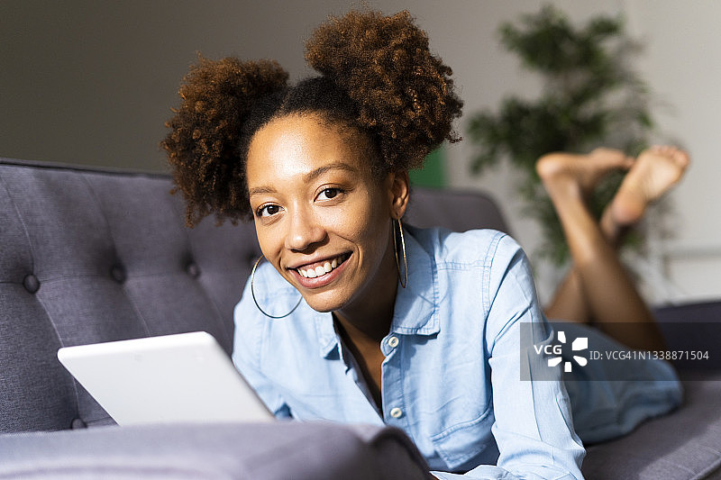 一个带着数码平板电脑微笑的非洲女人躺在客厅的沙发上图片素材