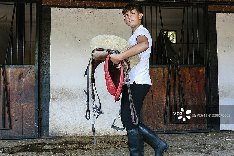 一个穿着骑士服装的少年。以马厩和谷仓为背景。青少年，乡村，村庄，马，骑手，和度假的概念。图片素材