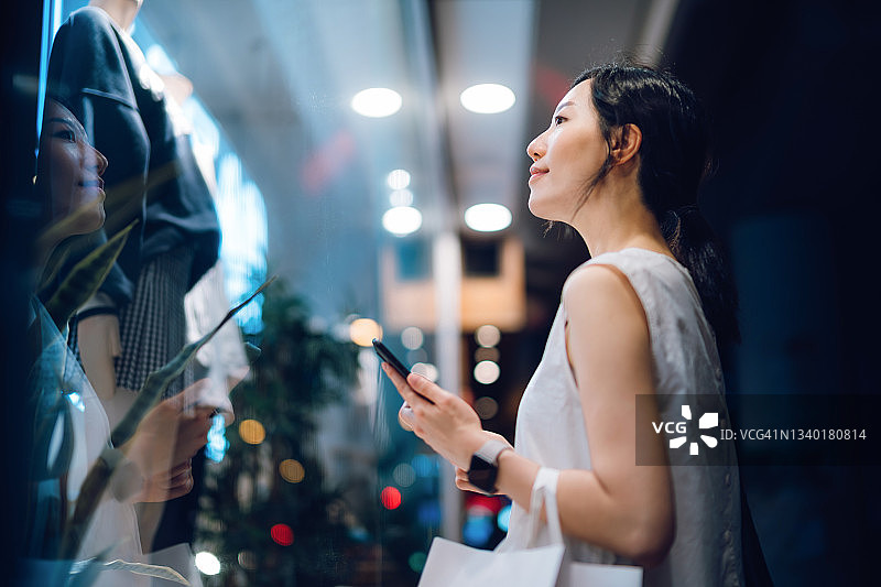 晚上，一名年轻的亚洲女子背着一个纸质购物袋，站在一家精品店外看着商店的橱窗，查看她的智能手机图片素材