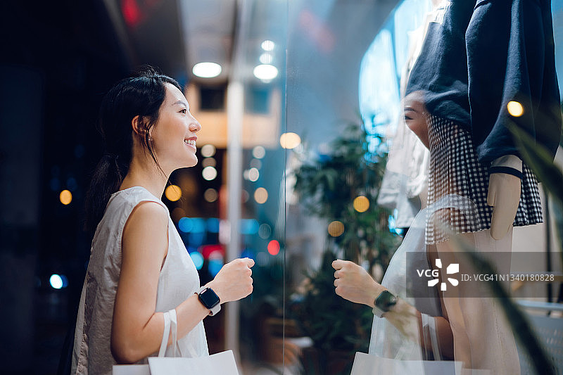 一个微笑的年轻亚洲女子背着一个纸购物袋，站在一家精品店外看着商店的橱窗，在晚上的城市图片素材