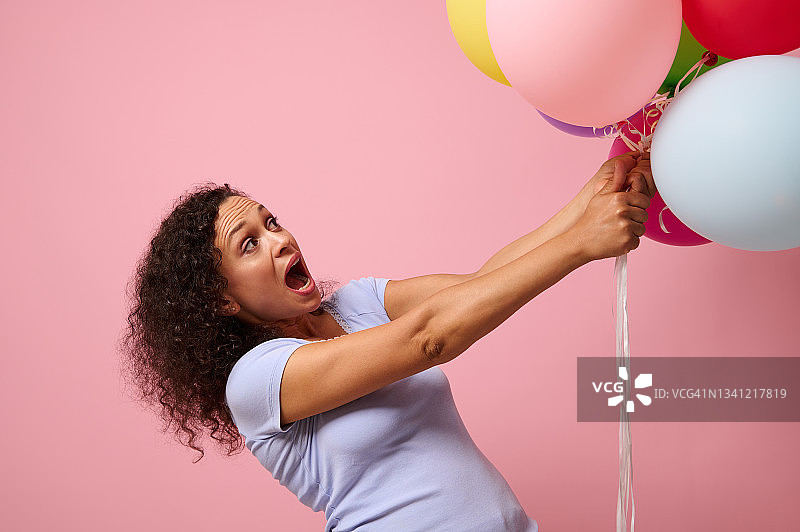 工作室为广告拍摄了一个惊讶的女人伸手去拿彩色气球，站在粉红色的背景下。庆祝、节日、周年纪念和礼物的概念。图片素材