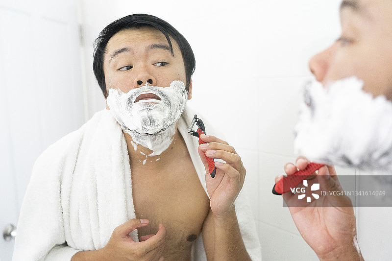 聪明的亚洲年轻人在浴室用白色泡沫刮胡须图片素材