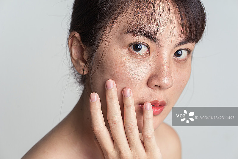 年轻的亚洲女性担心脸上有雀斑，用手轻轻触摸脸颊，应用护肤化妆品治疗。图片素材