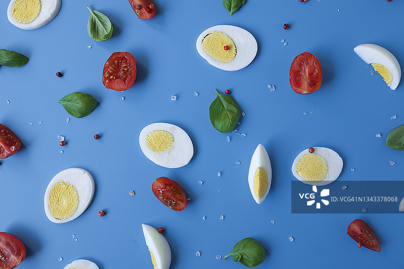将煮熟的鸡蛋、西红柿、罗勒、盐和红辣椒平铺在蓝色背景上图片素材
