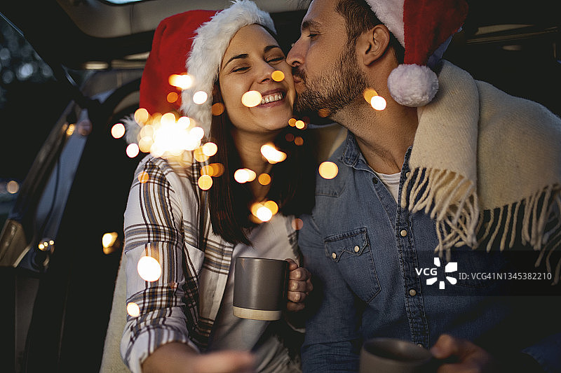 年轻的情侣在圣诞节坐在车里，打开后备箱，戴着圣诞老人的帽子，燃烧着烟花图片素材