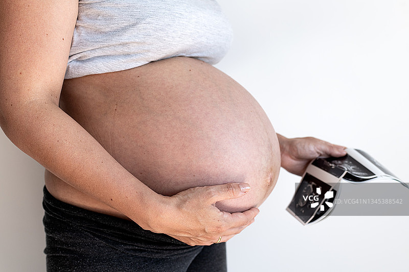 一个非常怀孕的女人腹部的右侧面特写。她穿着衬衫，左手拿着婴儿的超声波图。图片素材