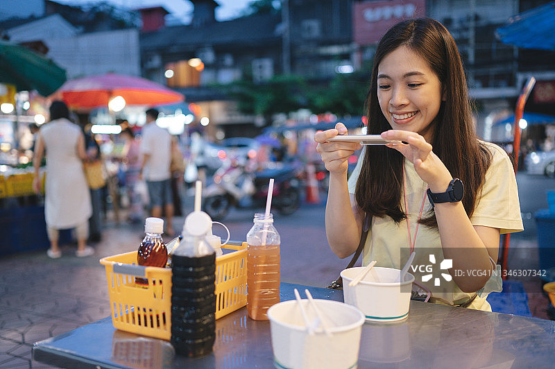 年轻的z世代，当地亚洲人周末快乐，使用智能手机5G互联网社交媒体直播，在泰国曼谷的唐人街吃鱼丸面街头小吃图片素材