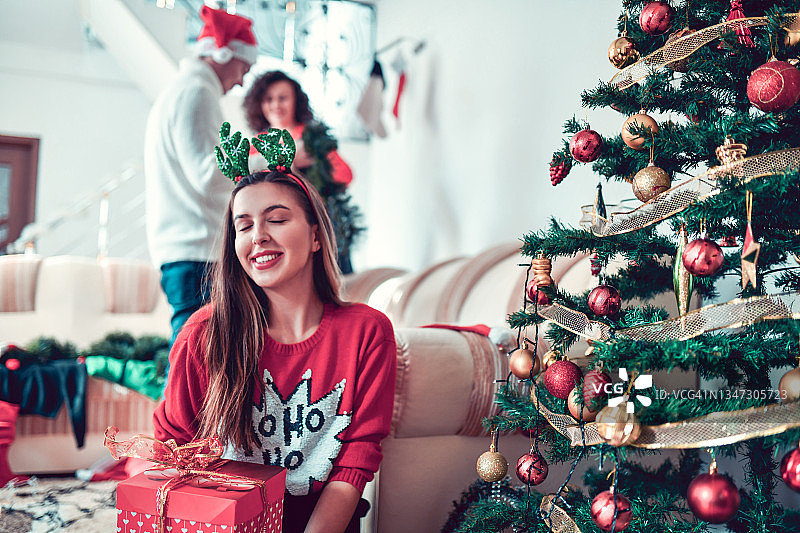 微笑的女性闭上眼睛和圣诞鹿角准备打开礼物树下图片素材