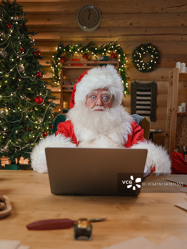 圣诞老人带着惊讶的表情看着镜头，我们看到他在他装饰过的办公室里，面前放着笔记本电脑图片素材