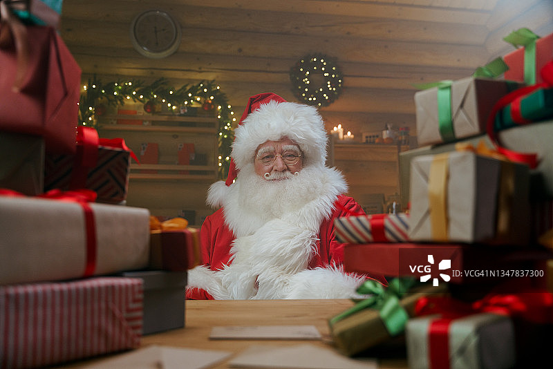 在温暖的灯光下，圣诞老人坐在他装饰华丽的办公室里的桌子旁，后面是一捆捆的礼品盒图片素材