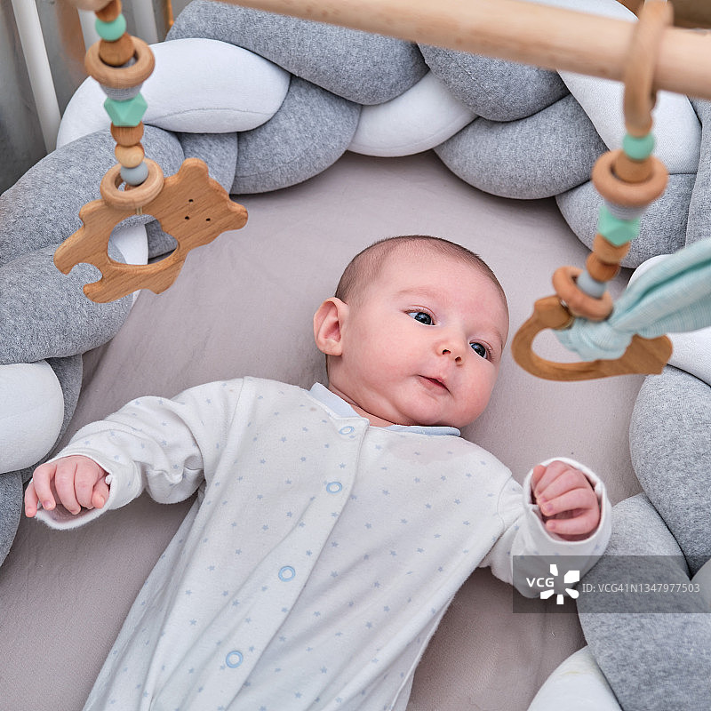 微笑的小男孩在玩挂在婴儿床上方的木制手机。快乐的孩子在床上玩玩具图片素材