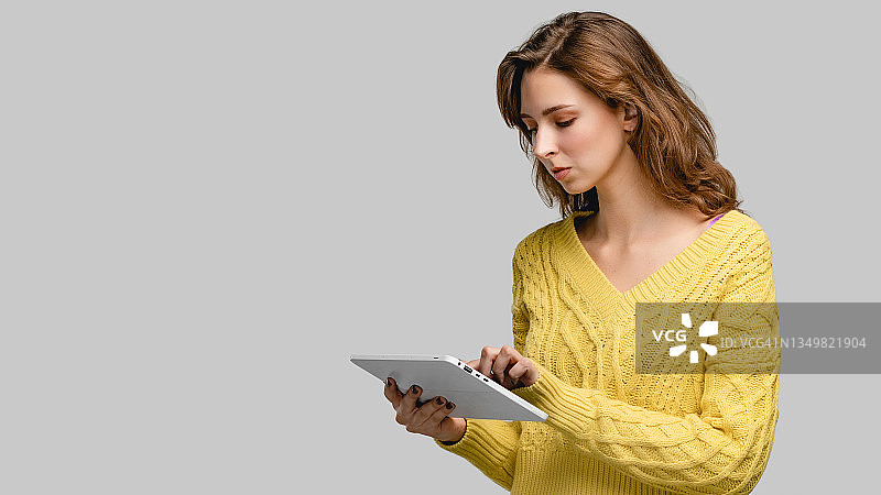 使用数码平板电脑在线工作的人。画室女子肖像女孩穿着黄色毛衣。概念照片与人在灰色背景和复制空间。图片素材