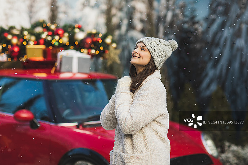积极的女人在羊毛手套和毛皮夹克站在旁边的汽车与绿色的圣诞树和礼物在顶部。图片素材
