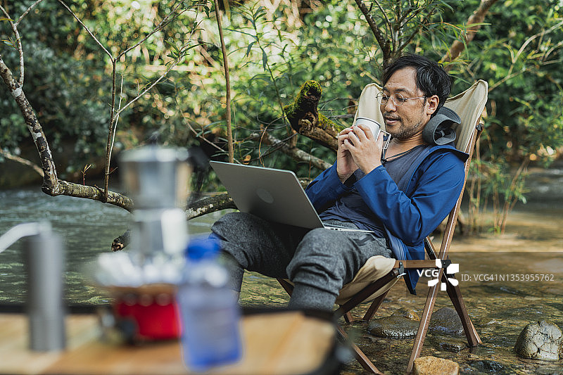 资深亚洲人坐在户外的椅子享受和回复电子邮件或聊天的笔记本电脑在流。自由职业者男性工作远程在茂密的森林在早上。年轻人在山间小溪边休息图片素材