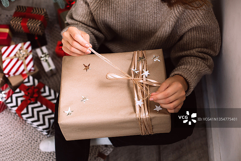 在圣诞树下，一名女子坐在编织的地毯上，打开印有银星的纸制礼品盒。新年庆祝概念。前视图。可持续的生活方式图片素材