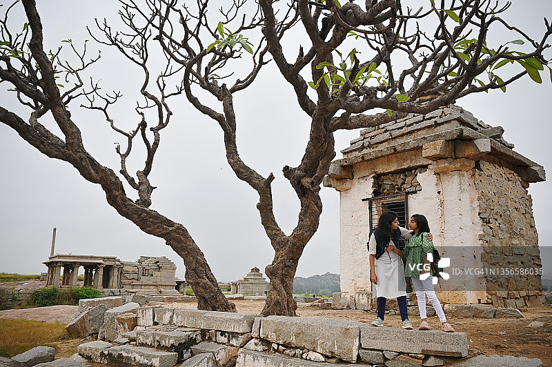 在卡纳塔克邦的罕比，在古老废墟的贫瘠景观中，母亲抱着女儿站在一个小哈努曼神殿和一棵树旁图片素材