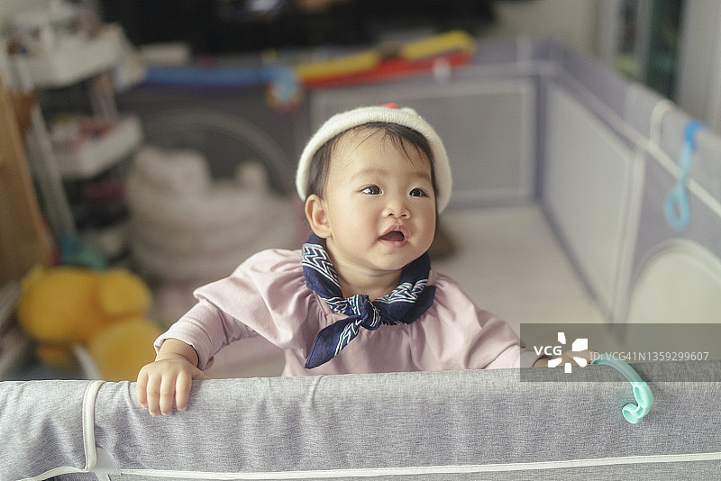 亚洲女婴面部表情和积极情绪穿上冬装，站在客厅内的婴儿围栏上过家庭生活图片素材