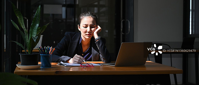 一位疲惫的年轻女商人正拿着笔记本电脑坐在办公桌旁。图片素材