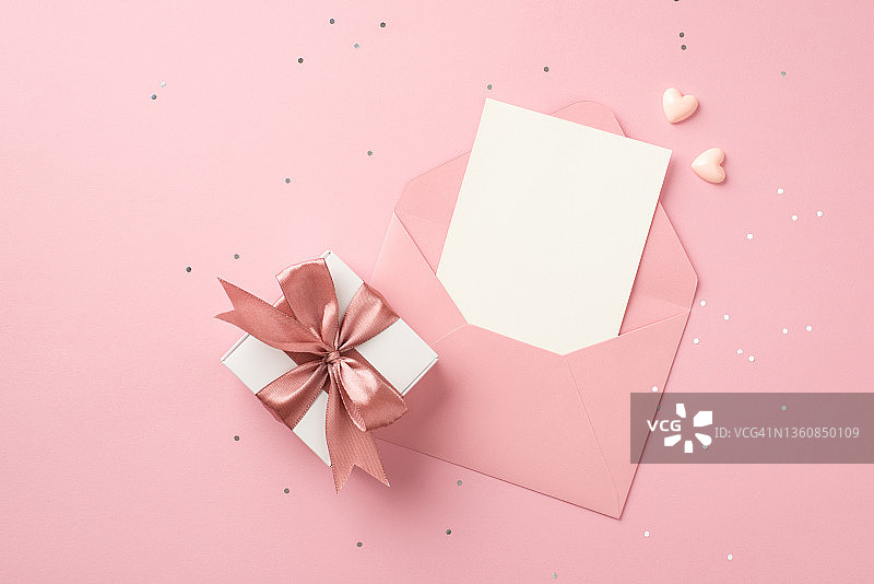 俯视图照片白色礼盒与粉红色丝带蝴蝶结小心脏打开粉红色信封与纸卡片和闪亮的亮片在孤立的粉彩粉红色背景与空白空间图片素材