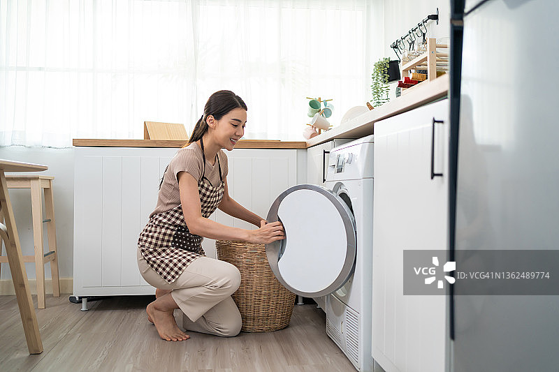 亚洲美女把脏衣服放在家里的洗衣机里。漂亮的女孩穿着围裙坐在地板上，高兴地把衣服放进家里的洗衣机里洗。居住房屋保持概念。图片素材