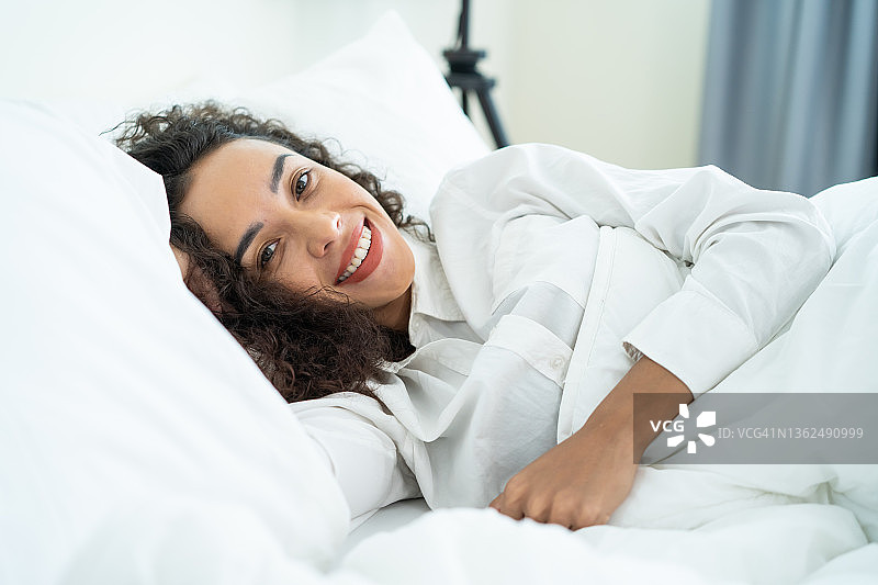 拉丁美人的肖像穿着睡衣微笑，看着相机。有魅力的年轻女性感到新鲜和快乐，从床上早起在家里早上的卧室。健康的生活方式的概念。图片素材