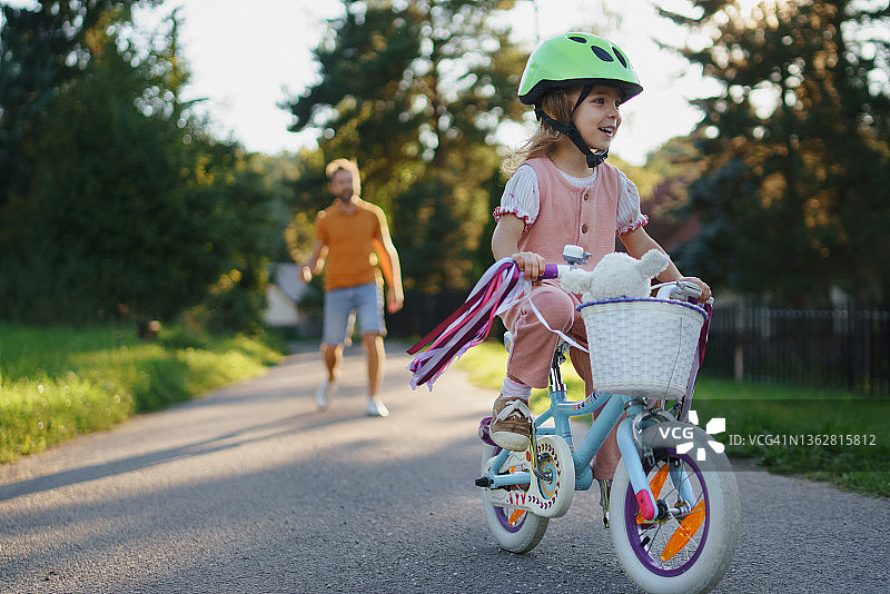 快乐的小女孩在公园里学习骑自行车，她的父亲在后面。图片素材