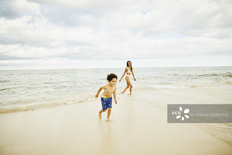 在热带海滩度假时，微笑的女人和儿子在水里玩耍的宽镜头图片素材