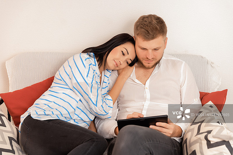 一个白人夫妇拥抱和花时间一起在沙发上看平板电脑的肖像图片素材