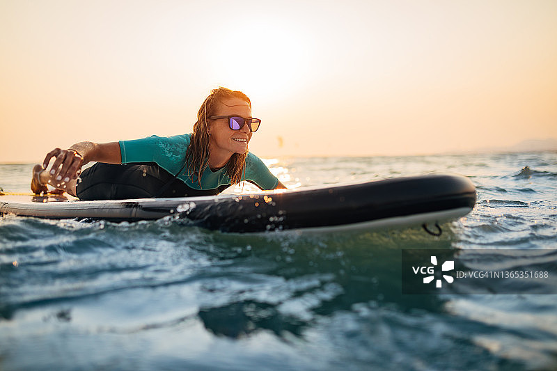 微笑的年轻女子戴着太阳镜躺在一个SUP板在大海和享受海浪。图片素材