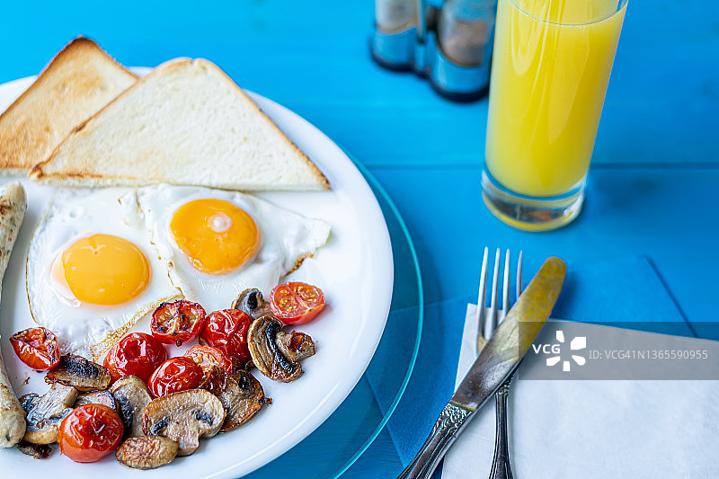 早餐有煎蛋、烤香肠、炸蘑菇、圣女果和酥脆的吐司，放在蓝色木制桌子上的白色盘子里，还有一杯橙汁。图片素材
