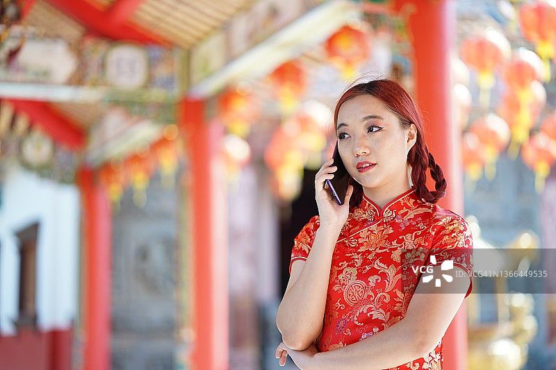 肖像美丽的亚洲年轻女子穿着红色的中国传统旗袍装饰和使用智能手机。图片素材