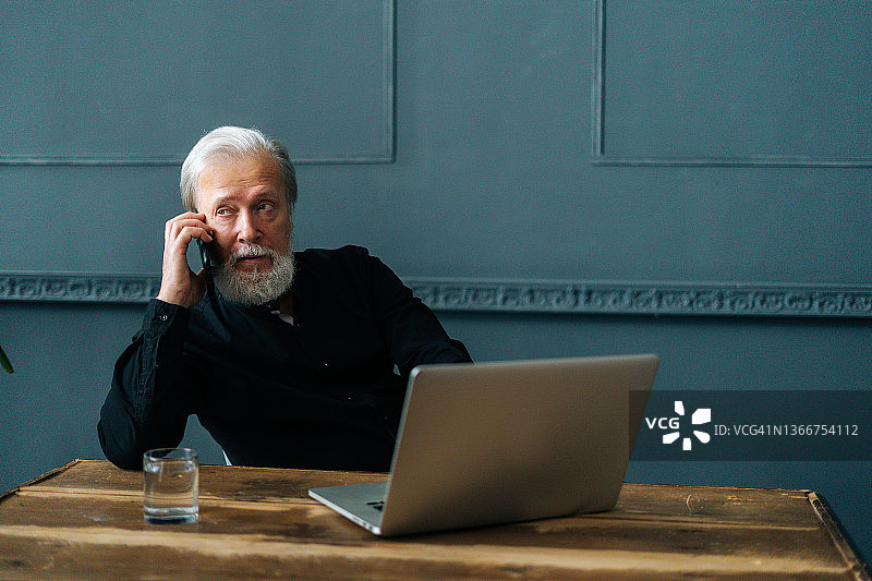 肖像严肃的灰色头发的高级成人商业人通话的移动电话坐在木桌与笔记本电脑。图片素材