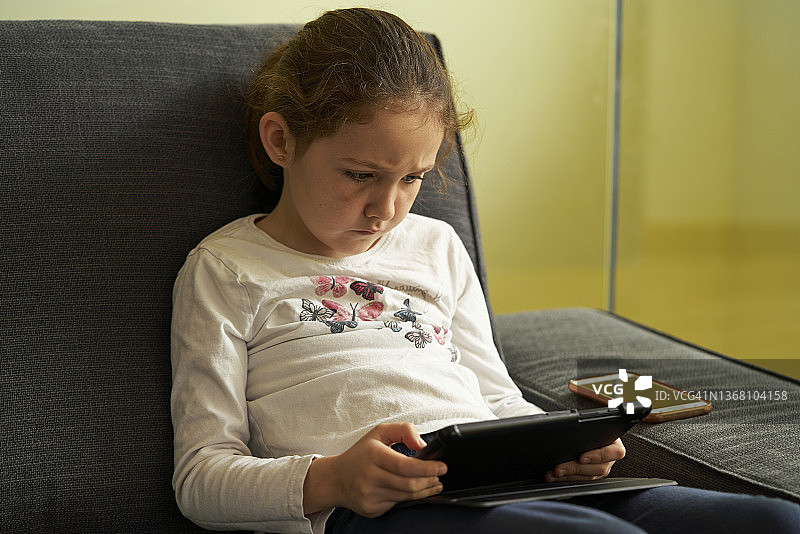 女孩坐在家里的沙发上玩平板电脑。图片素材