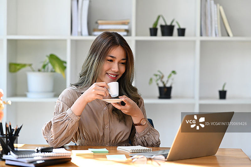 体贴的亚洲商业妇女喝热咖啡和阅读电子邮件的笔记本电脑。图片素材