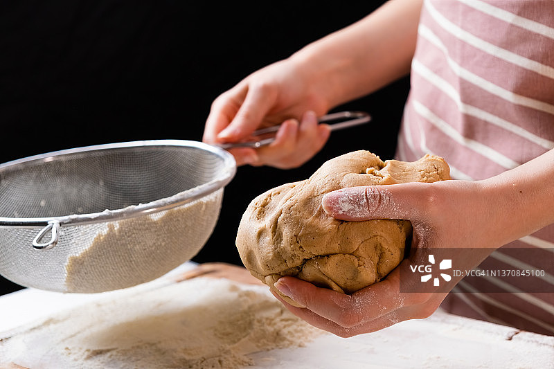 特写镜头:在黑色的背景上，一名妇女通过筛子将小麦粉筛入搅拌碗中，为蛋糕和面包店准备面团。揉面团做自制糕点图片素材
