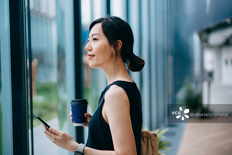 年轻成功的亚洲女商人，拿着智能手机，站在窗前，向前看，在一个现代的办公空间。她穿着时髦的便装上班。规划她的事业和未来。工作场所的女性。性别平等。忙碌的生意图片素材