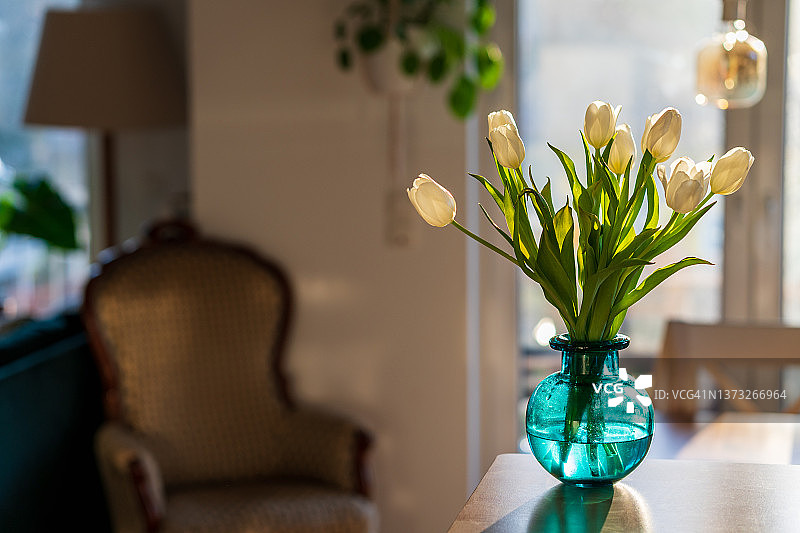 清晨明亮的阳光下，蓝色绿色的玻璃花瓶里插着一束白色郁金香。现代的室内设计，清新的春天鲜花在舒适的客厅。家居设计，装饰和舒适的概念。水平的。图片素材