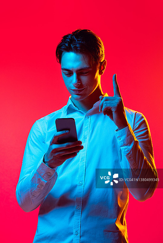 年轻男子的肖像，学生看着手机和竖起的手指孤立在红色的工作室背景蓝色霓虹灯图片素材