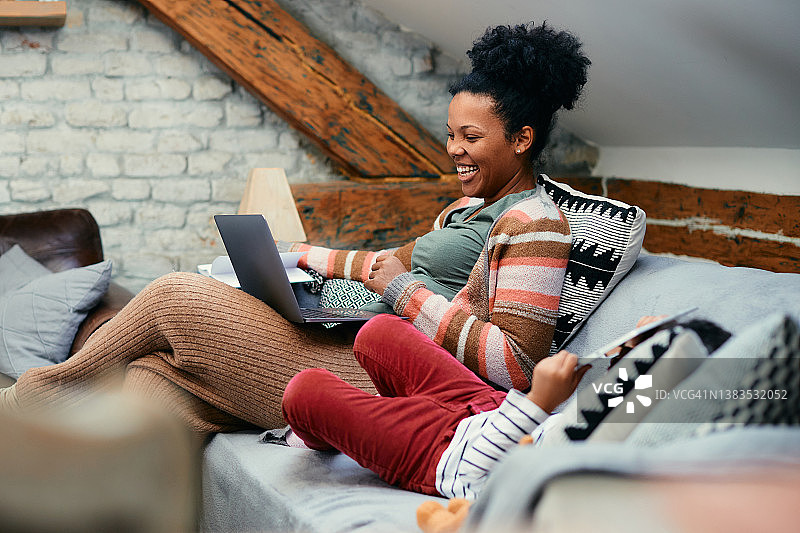 快乐的黑人母亲一边和女儿在一起一边笑，一边在家里用笔记本电脑工作。图片素材