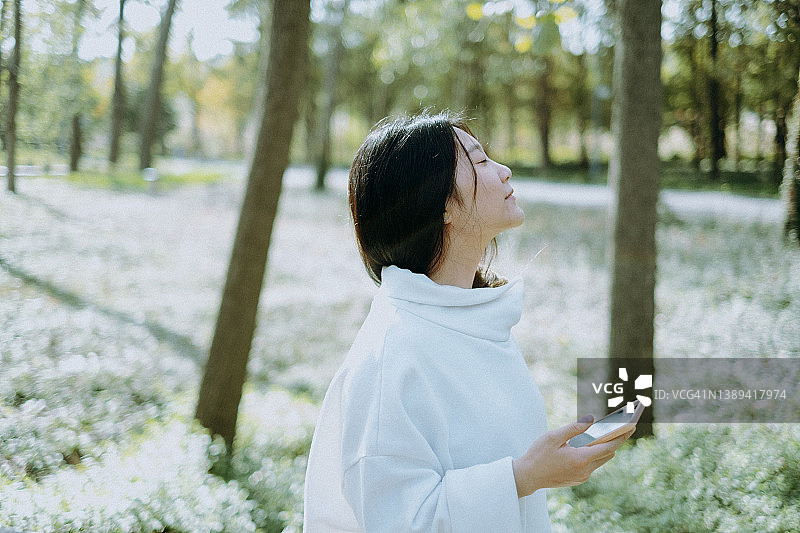 年轻的亚洲妇女使用手机在公园享受早晨温暖的阳光图片素材