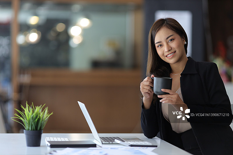 年轻的亚洲女商人在办公室工作时喝咖啡休息。看相机。图片素材