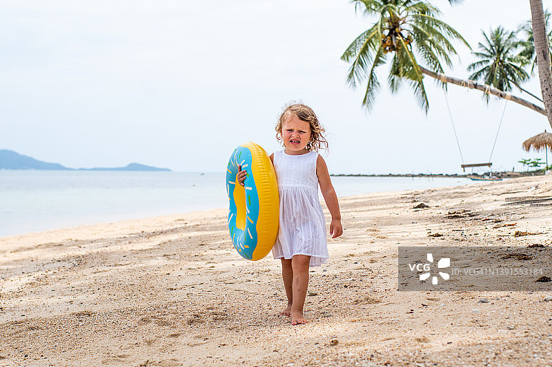 一个穿着白色连衣裙的卷发小女孩在沙滩上散步，笑着。孩子拿着一个充气的游泳圈。和孩子一起旅行和度假图片素材